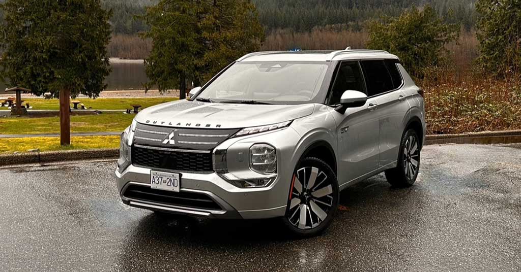 2026-Outlander-PHEV - Etobicoke Voice Front-three-quarter view of a silver Mitsubishi Outlander SUV parked on a wet pavement with trees and a lake in the background, daytime outdoor scene.